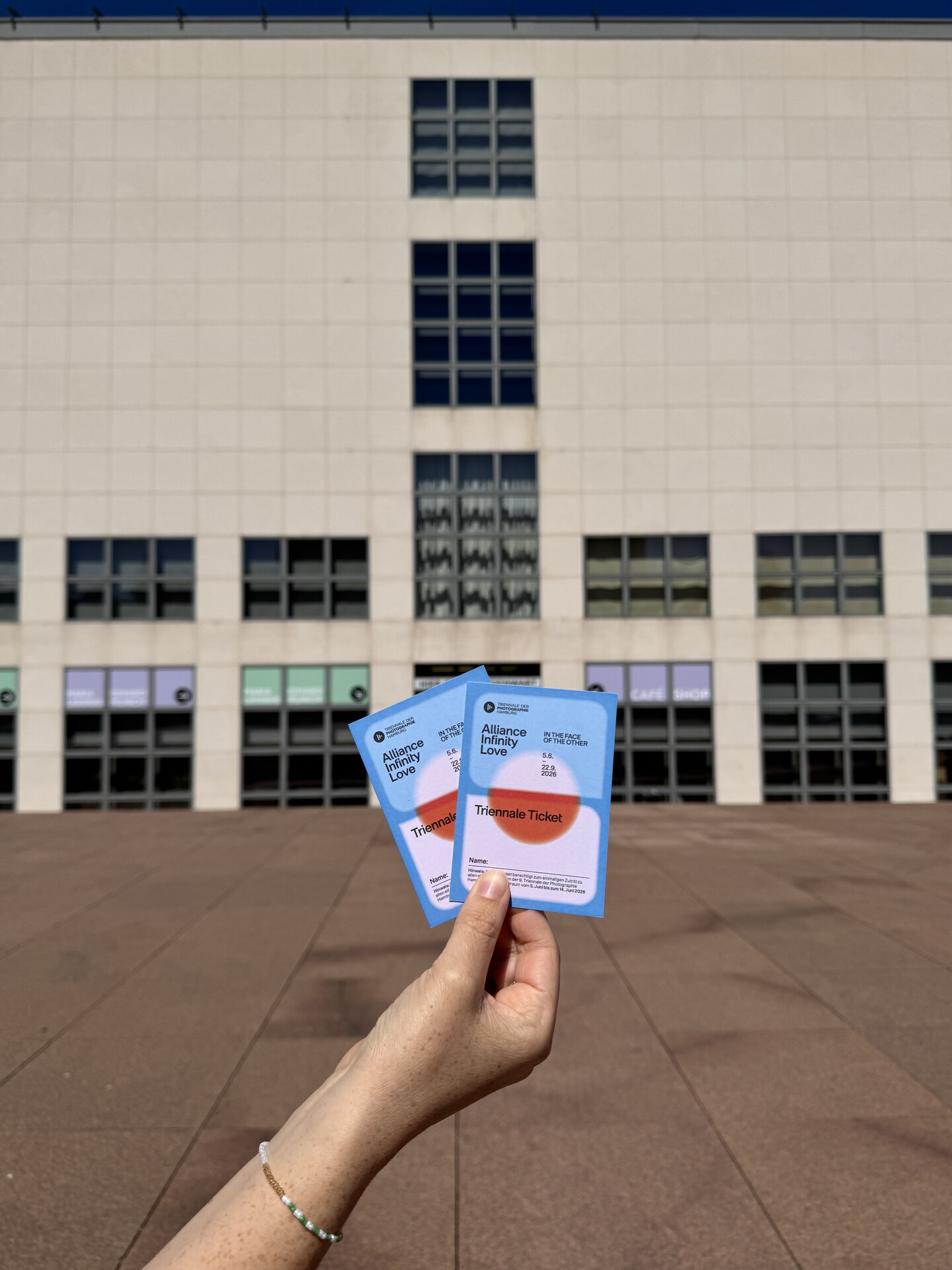 Hand hält zwei Tickets vor einem Gebäude mit vielen Fenstern