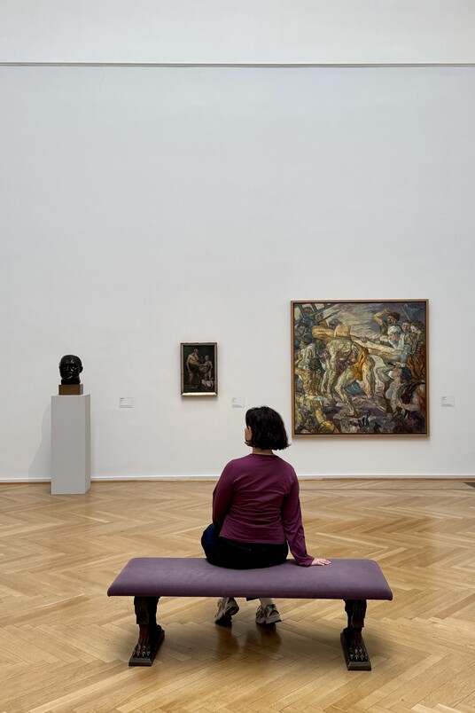 Person sitzt auf Bank in hellem Galerieraum mit Kunstwerken von Max Beckmann